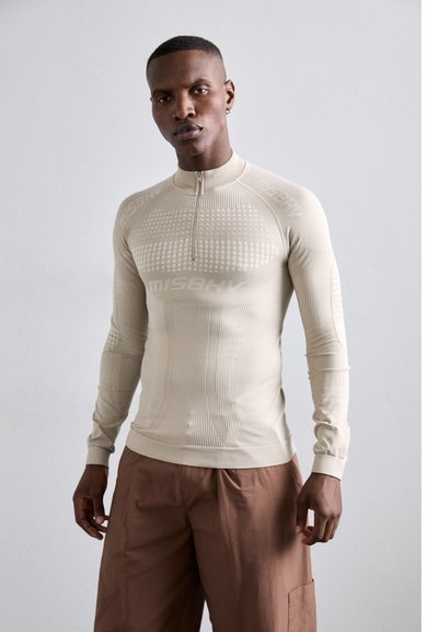 Haut de sport beige à manches longues avec un zip quart, un côtes texturé et des motifs à pois. Nom de la marque visible sur la poitrine et les épaules.
