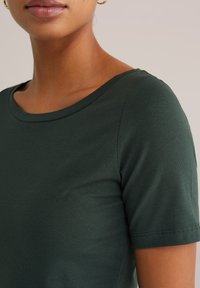 Tumši zaļš īsām piedurknēm tops ar apaļu kakla izgriezumu. Audums šķiet gluds un piegulošs, ar vieglu stiepšanos un kārtīgām šuvēm.