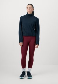 Mujer de pie vistiendo una camiseta de manga larga con cremallera en color navy, leggings burdeos, calcetines blancos y zapatillas de correr negras sobre un fondo liso.