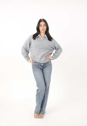 Pullover grigio a maglia con colletto alto, abbinato a jeans azzurri a vita alta. La modella si trova in piedi con le mani sui fianchi su uno sfondo bianco.
