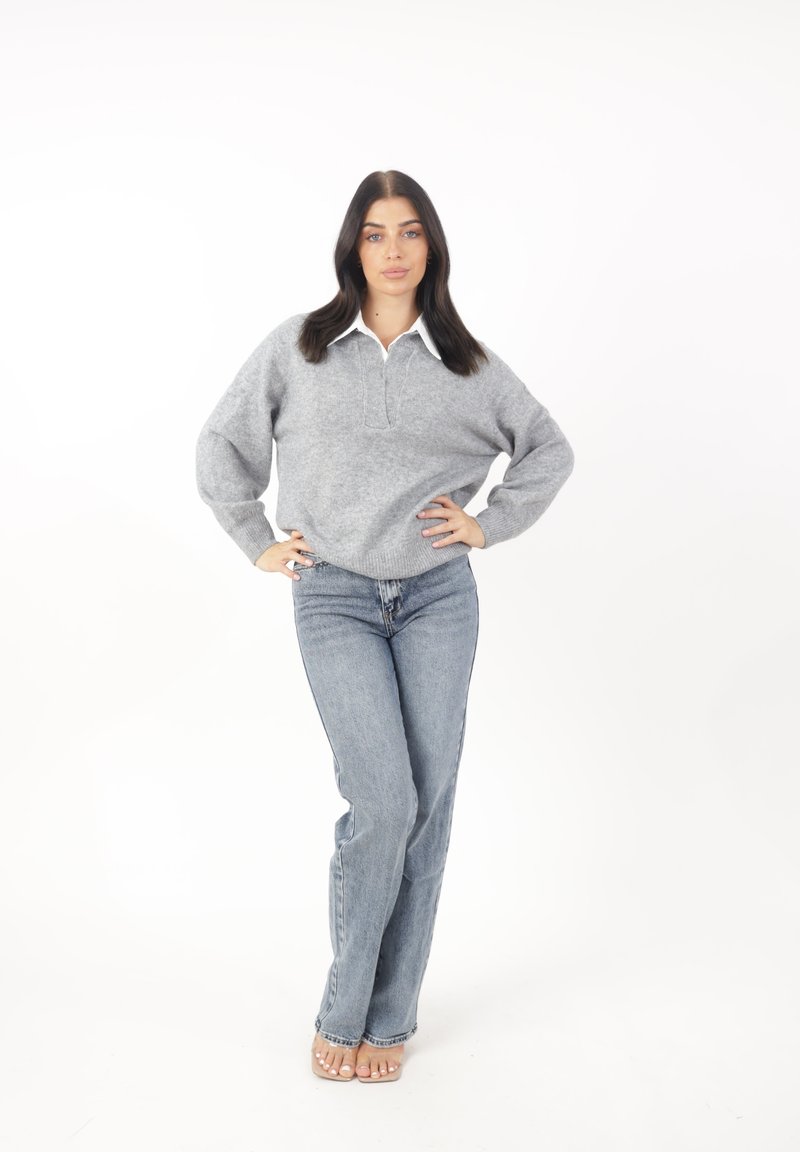Grijze gebreide pullover met een opstaande kraag, gecombineerd met lichtblauwe hoge taillejeans. Model staat met handen op de heupen tegen een witte achtergrond.