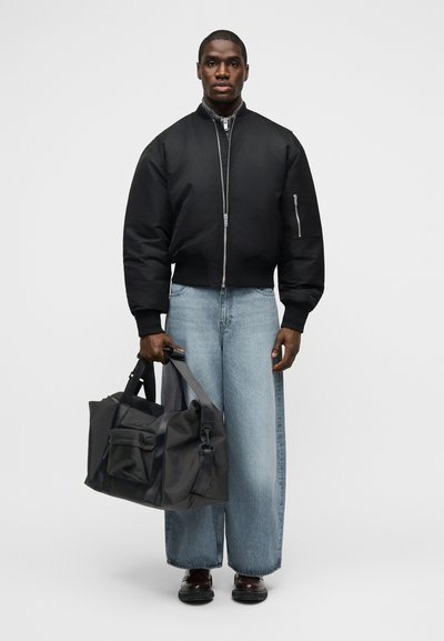 Άνδρας που φοράει μαύρο μπουφάν τύπου bomber και φαρδιά ανοιχτό μπλε τζιν παντελόνια, κρατώντας μεγάλη μαύρη τσάντα ταξιδιού με διπλές χειρολαβές μπροστά σε λευκό φόντο.