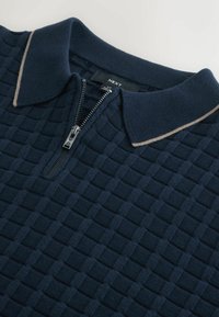 Polo shirt tricotat de culoare navy, cu un model texturat în grilaj, guler contrastant de bej deschis și închidere cu fermoar frontal. Eticheta brandului este vizibilă.