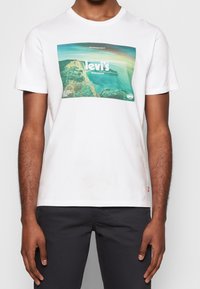 Camiseta de algodón blanca con un gran estampado gráfico colorido de una escena costera con el texto "Levi's Worldwide". Manga corta y cuello redondo.