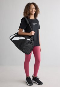 Sac de sport noir avec des rayures blanches, fabriqué en tissu durable. Le modèle porte un t-shirt noir avec un logo blanc, un legging rose et des baskets noires.