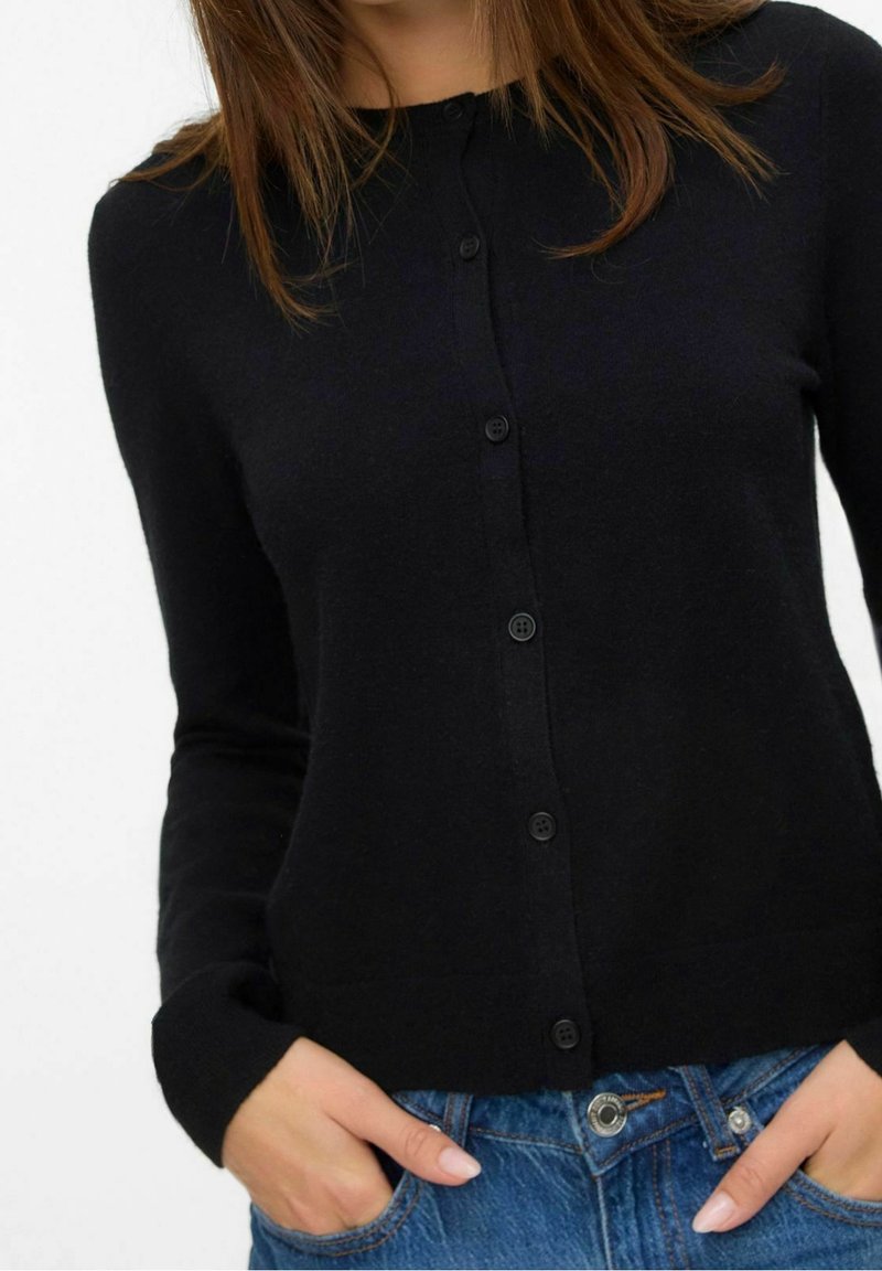 Femme portant un cardigan noir boutonné et un jean bleu avec les mains dans les poches, cadrée du cou aux hanches, fond uni.