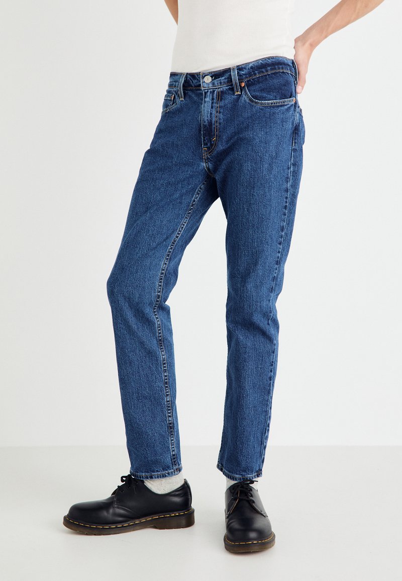 Blå denim jeans har et rett ben-design med subtil falming, fem lommer og kontrasterende sømmer. Båret med svarte sko.