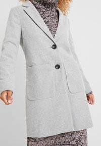 Light gray wool coat with a tailored design, two front pockets, notched lapels, and two black buttons, worn over a patterned sweater dress.