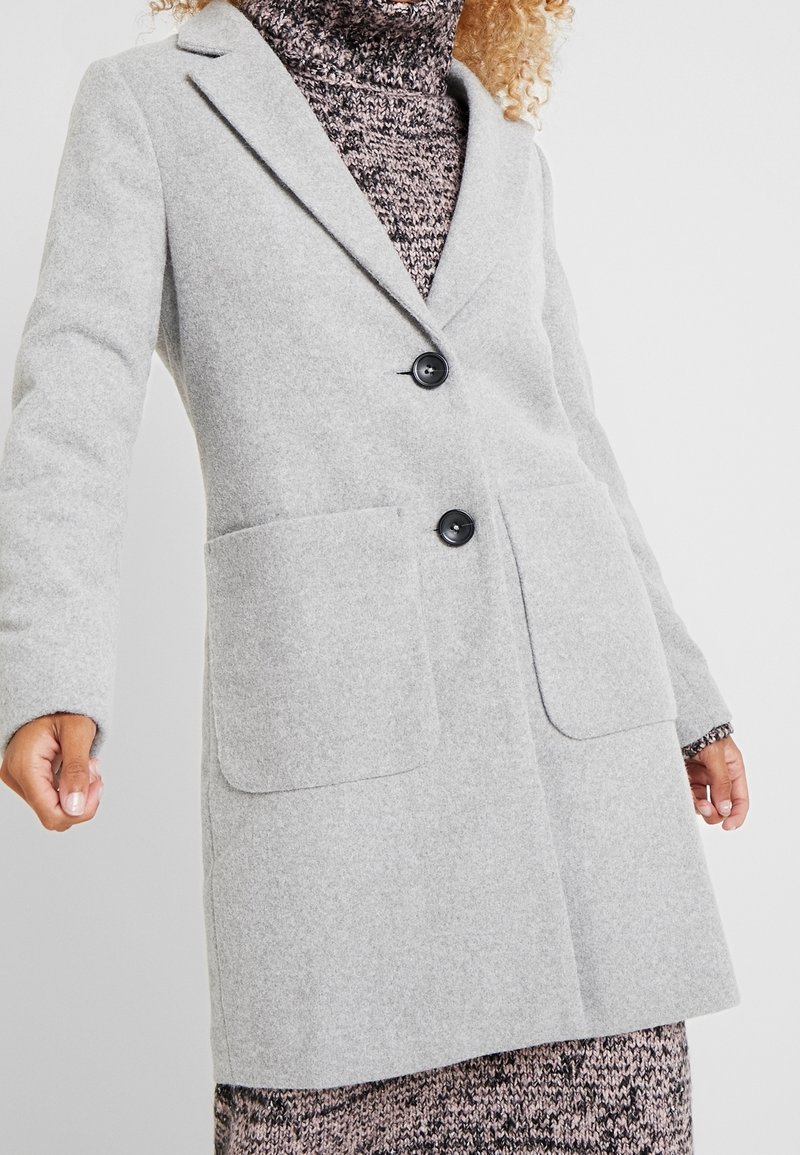 Light gray wool coat with a tailored design, two front pockets, notched lapels, and two black buttons, worn over a patterned sweater dress.