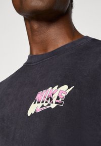 Čierne bavlnené tričko s grafikou loga "NIKE" v ružovej a bielej farbe, umiestnené diagonálne, s okrúhlym výstrihom a mäkkou textúrou.