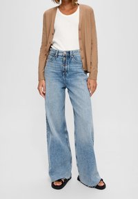 Beige kofta, vit t-shirt och ljusblå vida jeans med hög midja och knappgylf. Svarta sandaler med tjocka remmar och texturerad sula.