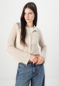 Beige kantgebreide cardigan met een kraag, knoopsluiting en uitlopende mouwen, gecombineerd met hoog-taille blauwe jeans.