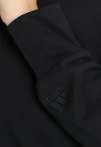 Sudadera de algodón negra con un acabado texturizado, que cuenta con un sutil logo de Adidas en relieve en el puño y un cierre frontal con cremallera.