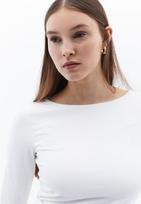 Maglietta bianca a maniche lunghe con un design aderente, texture liscia e scollatura ampia. Il modello indossa orecchini a cerchio dorati. Sfondo semplice.