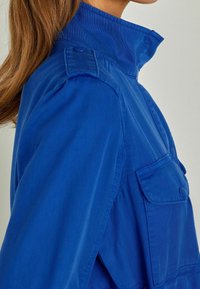 Woman’s upper torso wearing bright blue jacket with high collar, buttoned shoulder strap, and buttoned chest pocket, shown from side.