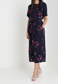 Vestito a fiori blu navy con maniche corte e vita annodata, caratterizzato da motivi floreali rossi e bianchi. Abbinato a sandali argentati con tacco alto.