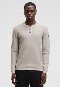 Beige Langarmpullover mit gerippter Struktur, runder Halslinie, einer drei-Knopf-Leiste und einem kleinen schwarzen Logo-Patch am Ärmel.