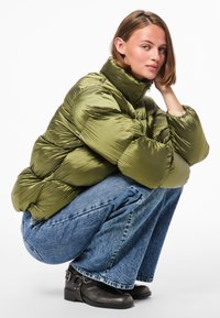 Olive green puffer jacket with a high collar, ribbed texture, and puffy silhouette, worn over wide-leg denim jeans and black ankle boots.