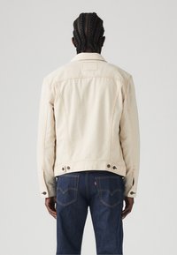Beige spijkerjasje met oranje stiksels, met knoopsluitingen bij de mouwen en een rugpandontwerp, gecombineerd met donkerblauwe jeans met achterzakken.