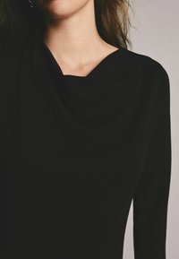 Must musträkkidega must topp, millel on drapeeritud kaelus, sujuva kangaga, rõhutades salendavat ja minimalistlikku siluetti.