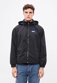 Jachetă neagră ușoară cu glugă, accente albastre, închidere cu fermoar, manșete elastice și o patch cu logo în față. Textură netedă, croială casual.