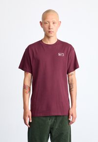 Homem com tatuagens, vestido com uma t-shirt Nike cor vinho e calças verdes, de pé contra um fundo branco simples, com uma expressão neutra.