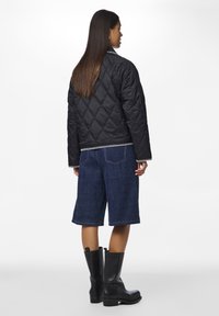 Giacca nera trapuntata con motivo a rombi, abbinata a pantaloni shorts in denim. Il modello indossa stivali alti neri in pelle e una camicia a righe sotto.