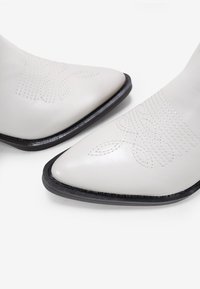 Stivaletti alla caviglia in pelle bianca con punta appuntita, caratterizzati da dettagli ricamati e una suola in gomma nera. Texture liscia e design elegante.