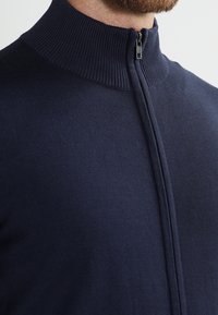 Marinblå zip-up tröja med hög krage, tillverkad av mjukt material. Har ribbade detaljer runt kragen och en strömlinjeformad design.