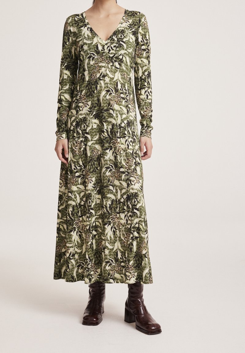 Femme portant une robe maxi à manches longues avec un col en V et un motif floral vert et beige, associée à des bottines à talons marron foncé.
