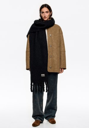 Czarny szalik z frędzlami, wykonany z teksturowanego materiału. Nosi się go na beżowej, oversize'owej kurtce z dużymi guzikami i luźnych dżinsowych spodniach.