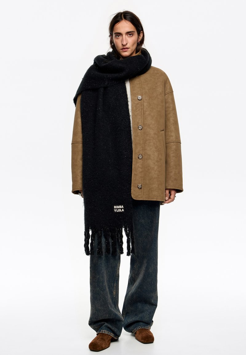 Bufanda negra con flecos, hecha de un material texturizado. Lleva puesta sobre una chaqueta oversized color canela con grandes botones y pantalones de denim holgados.