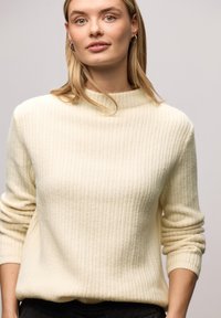 Kremowy sweter w prążki z wysokim kołnierzem i podwiniętymi mankietami, wykonany z miękkiej tkaniny, oferujący luźne dopasowanie.