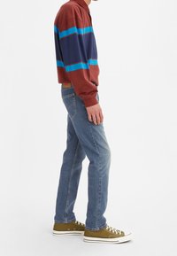 Person som bär blå jeans, olivgröna sneakers och en vinröd långärmad tröja med horisontella ränder i marinblått och blått, står på sidan.