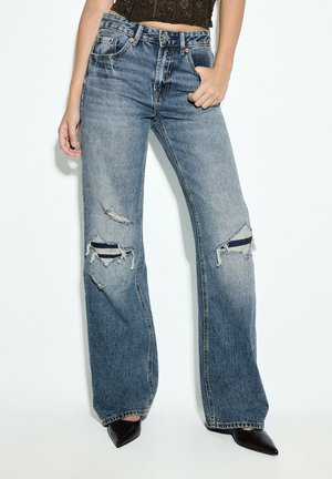 Kvinde iført højtaljede, vide blå jeans med revnede knæ og sorte spidse sko, stående mod en hvid baggrund.