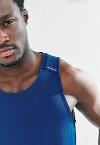 Niebieska sportowa koszulka na ramiączkach z teksturowaną tkaniną, szerokimi otworami na ramiona oraz odblaskowym akcentem na ramieniu. Zbliżenie na obszar ramienia.