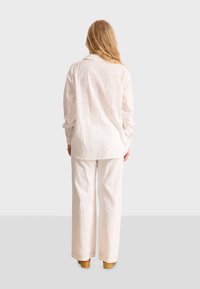 Camisa blanca con bordado de ojales y pantalones de pierna ancha, que presentan intrincados patrones cuadrados y una superficie texturada. El calzado son zuecos marrón claro.