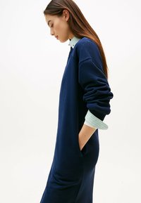 Navyblaues Sweatshirt-Kleid mit langen Ärmeln und Bündchen an den Handgelenken. Verfügt über eine weiche Textur und eine Tasche an der Seite. Über einem gestreiften Hemd getragen.