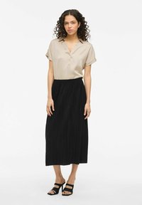 Camicia satin beige con scollo a V e maniche corte arrotolate, abbinata a una gonna midi nera plissettata. Sandali con tacco a strappo neri.