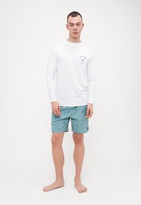 Hvit langermet rash guard med blå logo på brystet, kombinert med lyseblå svømmebukser med palmeprint.
