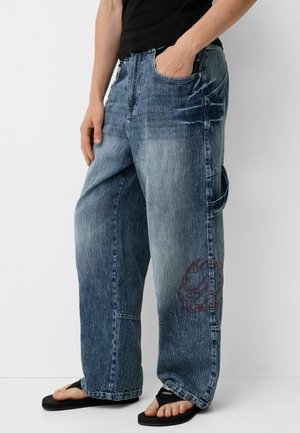 Person trägt locker sitzende, ausgewaschene blaue Jeans mit seitlicher Tasche und Blumenstickerei, kombiniert mit schwarzen Flip-Flops und schwarzem Oberteil.