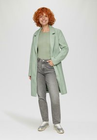 Mintgroene wollen jas met een gelapte kraag over een lichtgroene geribde top, gecombineerd met grijze jeans en metallic sneakers.