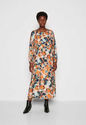 Γυναίκα που στέκεται σε ένα μακρύ, μακρυμάνικο φόρεμα με σχέδιο πορτοκαλί, μπλε και άσπρο σε αφηρημένο στυλ, φορώντας μαύρες μποτάκια μέχρι τον αστράγαλο, σε απλό φόντο.