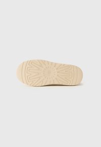 Semelle de chaussure beige avec un motif de semelle géométrique et le logo "UGG" en relief au centre, présentée sur un fond blanc.