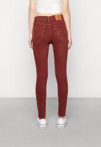 Jeans skiny burdeos de cintura alta, con dos bolsillos traseros, costuras en contraste y un parche de cuero marrón en la cintura.