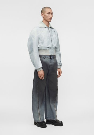 Άνδρας που φοράει ανοιχτό γκρι κοντή μπομπέρ ζακέτα, φαρδιά ξεθωριασμένα σκούρα τζιν και μαύρα ογκώδη παπούτσια σε απλό φόντο.