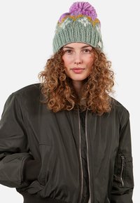 Knit beanie in light green with a textured pattern, featuring a purple pom-pom and gold accents. Worn with a dark bomber jacket.