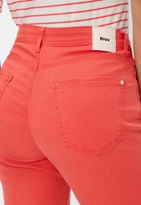 Koralfarbene Jeans mit hoher Taille, ausgestattet mit Gesäßtaschen und einem sichtbaren Markenlabel. Glatte Textur mit ausgeprägten Stitching-Details.