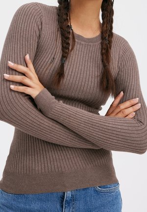 Personne aux cheveux tressés portant un pull marron côtelé à manches longues et un jean bleu, bras croisés sur la poitrine, devant un fond blanc.