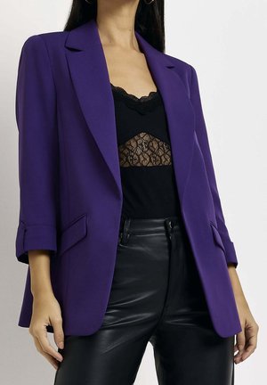 Mujer con una chaqueta morada sobre una blusa negra con encaje y pantalones de cuero negros, de pie frente a un fondo blanco liso.